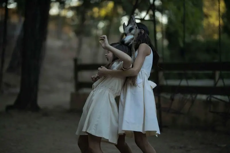 Two girls in "La casa del caracol", one wearing a wolf mask, interacting outdoors.